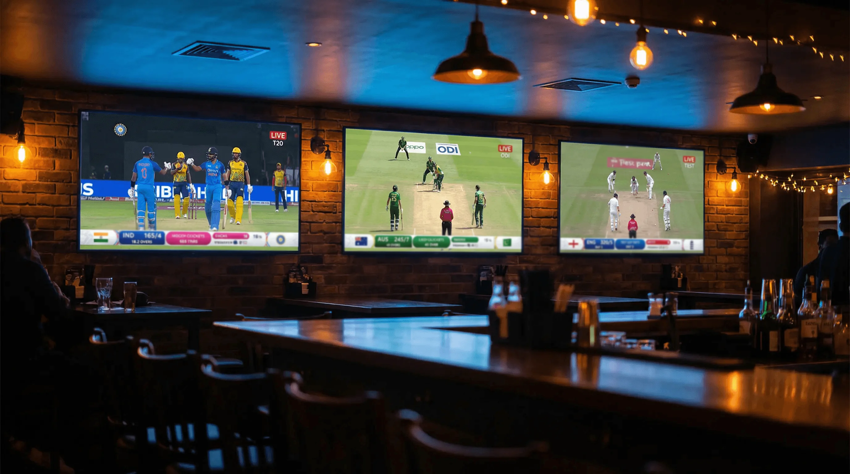Three cricket matches displayed on separate screens side by side in a sports bar setting with warm lighting