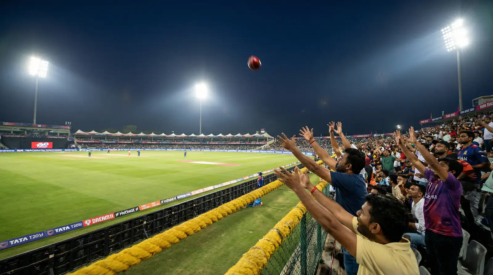 Cricket ball sailing high over the boundary rope into the crowd for a six during a T20 match at night